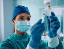 A female nurse wearing a face mask and gloves injecting medicine through an IV drip before a surgical procedure.