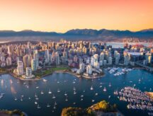 An aerial view of the downtown Vancouver skyline at sunset, with mountains visible in the background and sailboats in the foreground.
